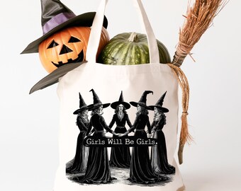 May include: A cream-colored tote bag with a black and white illustration of five witches holding hands, with the text "Girls Will Be Girls." The bag contains a carved orange pumpkin, a green pumpkin, and a broom. A black witch hat sits atop the orange pumpkin.