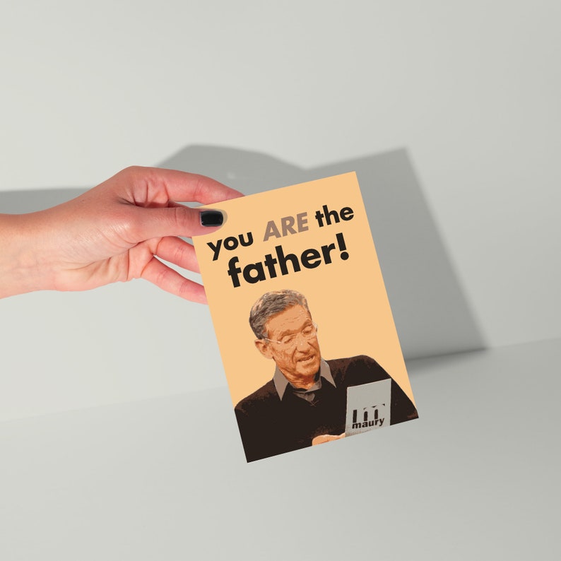 May include: A greeting card with the text "you ARE the father!" in black and a picture of a man. The card has a peach background and is held by a hand with black nail polish.