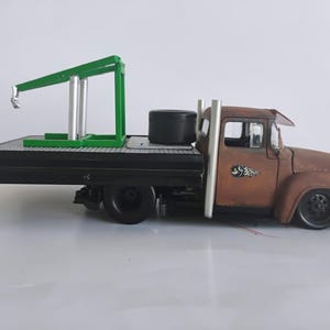 May include: A brown and black toy tow truck with a green crane. The truck has a flatbed and a black tire on the back. The truck has a rusted appearance and a decorative design on the door.