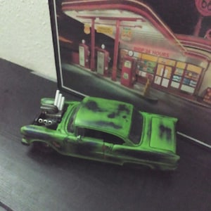 May include: A green and black toy car with an open hood, showcasing the engine. The car is positioned in front of a vintage gas station sign that reads "FAST SERVICE". The car has a weathered appearance.