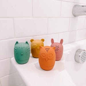 May include: Four colourful bath toys shaped like an owl, a cat, a bear, and a bunny. The toys are made of soft silicone and have a textured surface. They are sitting on a white bathroom sink.