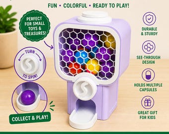 May include: A purple capsule dispenser with a white honeycomb-patterned window, filled with colorful capsules. The dispenser has a turn-to-spin mechanism and a collection tray. Text on the dispenser reads: "CAPSULE DISPENSER", "FUN • COLORFUL • READY TO PLAY!"