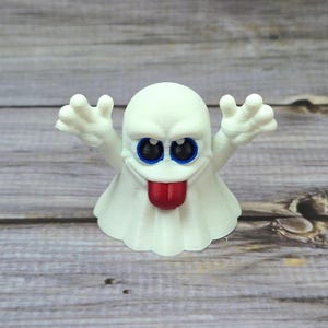 May include: A white 3D-printed ghost figurine with a round head, blue eyes, and a red tongue sticking out. The ghost has outstretched arms and a ruffled bottom edge. The figurine is on a wooden surface.