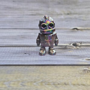 May include: A small, metallic robot figurine with a rainbow iridescent finish. The robot has a rounded head, large eye sockets, and a rectangular body. It stands on two feet and has small arms. The figurine is displayed on a wooden surface.