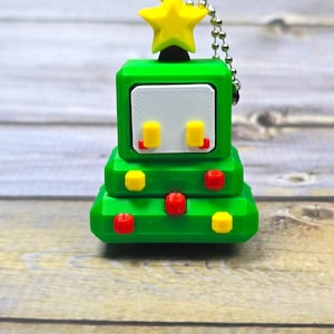 May include: A green, 3D-printed Christmas tree-shaped keychain with a yellow star on top. The tree has a white face with yellow and red accents, and is attached to a silver ball chain.