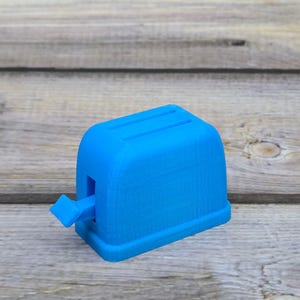 May include: A blue plastic toy toaster with a lever on the side. The toaster is on a wooden surface.