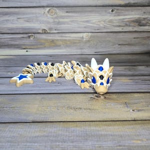 May include: A tan and white articulated dragon figurine with blue star and gem accents. The dragon has a long, segmented body and is posed on a wooden surface. The dragon's eyes and other details are painted in black.