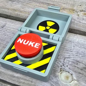 May include: A gray, hinged box with a red button labeled "NUKE" in white. Inside, a black and yellow hazard pattern surrounds the button. The box lid features a radiation symbol. A novelty item.