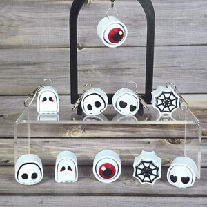 May include: A collection of Halloween-themed keychains displayed on a clear acrylic shelf and a black arch. The keychains feature designs like a ghost, an eyeball, a spiderweb, and a smiling face. The keychains are white with black and red accents.