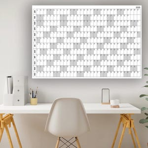 Könnte beinhalten: Ein weißer Trockenlöschkalender mit einem Raster aus Quadraten für jeden Tag des Jahres 2023. Der Kalender hängt an einer weißen Wand über einem weißen Schreibtisch mit einem weißen Stuhl, einer Pflanze und ein paar Büroartikeln.