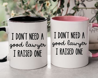May include: Two white coffee mugs with black lettering. The mug on the left has a black rim and handle. The mug on the right has a pink rim and handle. Both mugs say "I don't need a good lawyer I raised one."