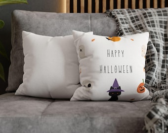 May include: Two white square throw pillows on a gray couch. One pillow has the words "HAPPY HALLOWEEN" printed in black, along with illustrations of a black cat, a pumpkin, ghosts, and a skull. A plaid blanket is draped in the background.