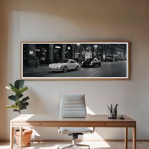 Könnte beinhalten: Ein panoramischer Schwarz-Weiß-Wandkunstdruck, der einen hellen Oldtimer-Sportwagen neben einem dunklen modernen Sportwagen auf einer nächtlichen Stadtstraße zeigt, gerahmt in einem dünnen Holzrahmen an einer schlichten Wand.