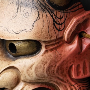 May include: Close-up of a traditional Japanese demon mask with a red and white painted face. The mask has a large, round eye with a gold-colored iris and a black pupil. The mask is decorated with black lines and swirls.