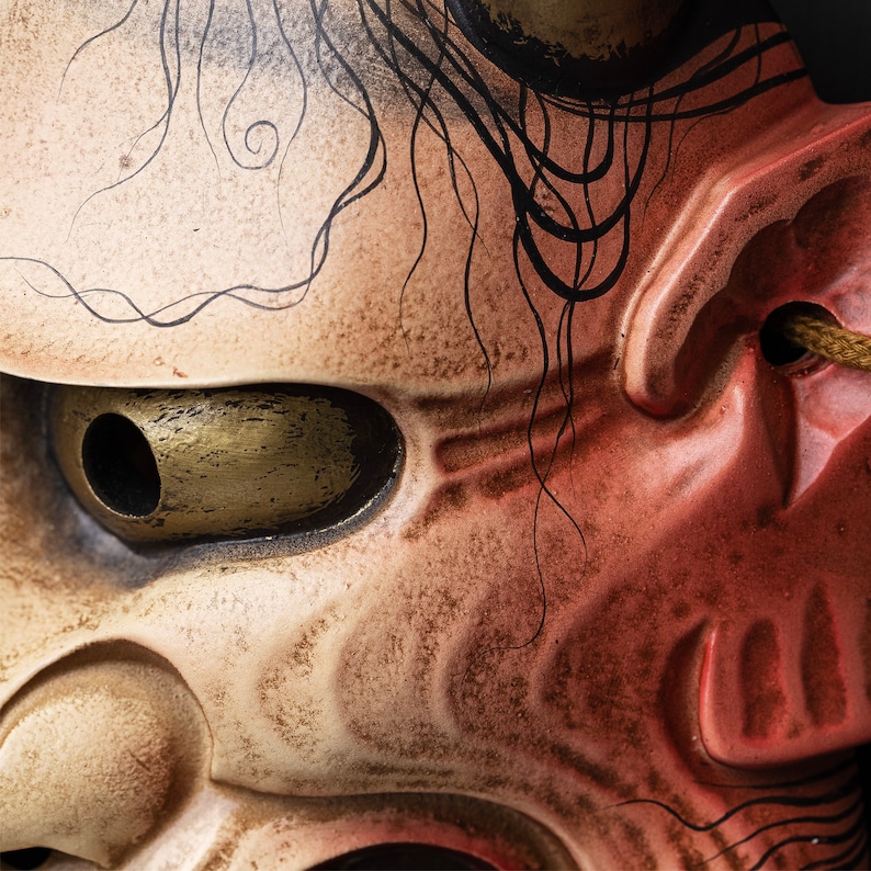 May include: Close-up of a traditional Japanese demon mask with a red and white painted face. The mask has a large, round eye with a gold-colored iris and a black pupil. The mask is decorated with black lines and swirls.