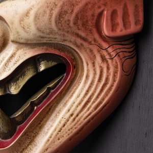 May include: A close-up of a traditional Japanese mask with a carved, detailed face. The mask is painted in shades of red, white, and gold, with a wide, open mouth and a fierce expression. The mask is made of wood and has a textured surface.