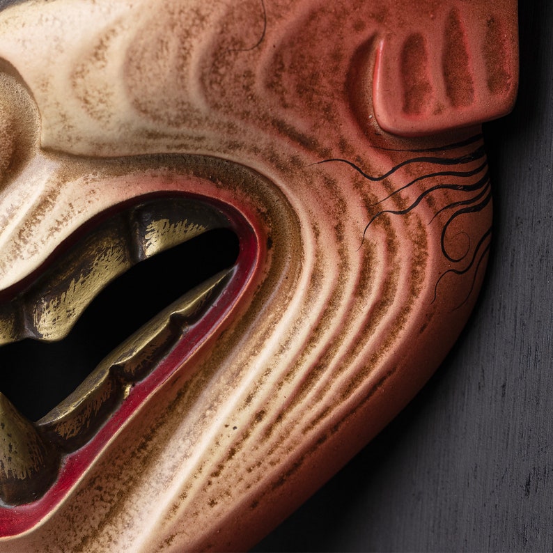 May include: A close-up of a traditional Japanese mask with a carved, detailed face. The mask is painted in shades of red, white, and gold, with a wide, open mouth and a fierce expression. The mask is made of wood and has a textured surface.