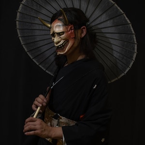 May include: A person wearing a black kimono and a traditional Japanese mask with red and gold accents. They are holding a black and white umbrella.