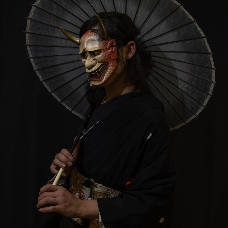 May include: A person wearing a black kimono and a traditional Japanese mask with red and gold accents. They are holding a black and white umbrella.