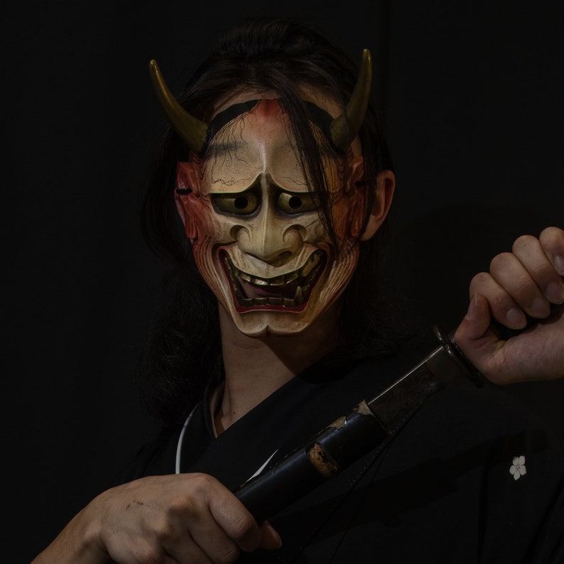 May include: A person wearing a traditional Japanese demon mask with red and gold accents, holding a black sword in their right hand. The mask has a fierce expression with sharp teeth and horns.