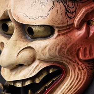 May include: A traditional Japanese mask with a fierce expression. The mask is made of wood and painted in red, white, and gold. It has a large, open mouth with sharp teeth and a long, flowing beard.