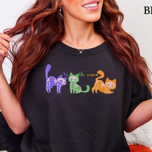 May include: Black t-shirt featuring three cartoon cats in purple, green, and orange, with the text "the season". A black and white mug with ghosts and stars is held in the hand. The word "BLACK" is printed in the upper right corner.