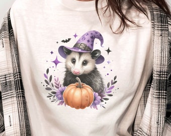 May include: Cream-colored t-shirt featuring a Halloween-themed graphic. The design includes a possum wearing a purple witch hat, holding a pumpkin, surrounded by purple flowers and stars. A black and white plaid shirt is worn over the t-shirt.