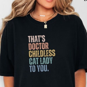 May include: Black t-shirt with the text "THAT'S DOCTOR CHILDLESS CAT LADY TO YOU." in a retro, multi-colored font. The shirt is a comfortable, oversized fit.