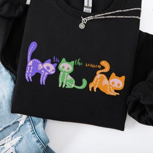 May include: Black sweatshirt featuring a colorful graphic of three skeleton cats with the text "'tis the season". The cats are purple, green, and orange. A silver necklace is draped across the sweatshirt.