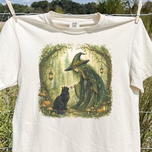 May include: An ivory short-sleeve t-shirt featuring a graphic print of a witch in a green cloak and pointed hat interacting with a black cat in a forest setting with hanging lanterns, mushrooms, and trees surrounding the central illustration.