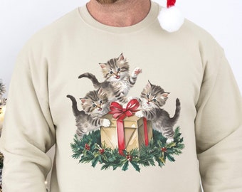 Könnte beinhalten: Sandfarbenes Sweatshirt mit festlichem Design. Es zeigt vier Kätzchen um eine Geschenkbox mit roter Schleife und Stechpalme. Das Wort "SAND" ist oben links aufgedruckt. Auf der linken Seite ist ein Weihnachtsbaum zu sehen.