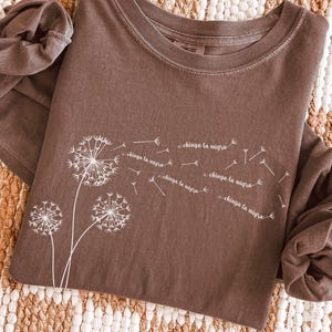 May include: Espresso-colored long-sleeve shirt featuring a dandelion design with the text "chinga la migra" in white. The shirt is made of a soft material and has a crew neck.