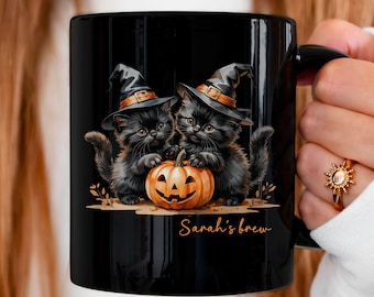 May include: Black ceramic mug featuring two black kittens wearing witch hats, flanking an orange jack-o'-lantern. The mug has the text "Sarah's brew" and is 11 oz.