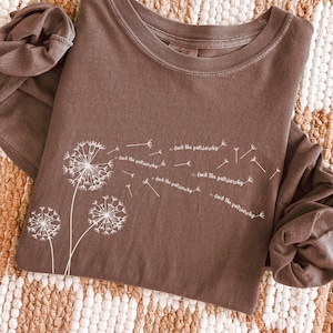 May include: Espresso-coloured long-sleeve shirt featuring a dandelion design and the text "fuck the patriarchy" in white. The shirt is made of a soft material and has a crew neck.