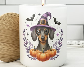 May include: A white candle with a lit wick, featuring a dachshund wearing a purple witch hat. The candle has a Halloween-themed design with pumpkins, bats, and stars. The candle is a cylindrical shape and has a wooden lid.