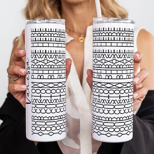 May include: Two white stainless steel tumblers with black geometric patterns and the name "Vanessa" in script. Each tumbler has a silver lid and a white straw. The tumblers are held by a person.