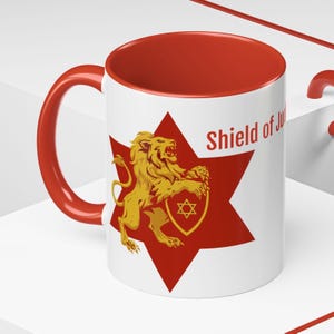 May include: White ceramic mug with a red interior and handle. The mug features a red star with a gold lion and a Star of David emblem. The words "Shield of Ju" are printed on the mug.