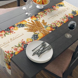 May include: A beige table runner with a colorful fruit and wheat design. The runner features peaches, grapes, and oranges, with Hebrew text. White plates and silverware are on the table, along with wine glasses and a bowl of fruit.