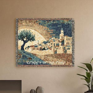 Puede incluir: Manta tejida con una imagen de estilo mosaico de una ciudad, un árbol y texto. La obra de arte utiliza tonos azules, marrones y beige. La manta tiene un borde con flecos y se exhibe en una pared neutra.