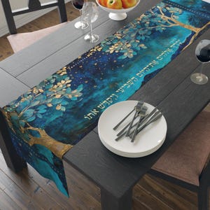 May include: A rectangular table runner featuring a deep blue and teal watercolor background with gold tree branches, leaves, and Hebrew text printed in gold. The runner is laid across a dark wooden table with a stack of white plates and silverware.