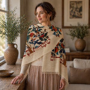 May include: A beige shawl with a colorful floral pattern in red, blue, and green. The shawl has a fringed edge and a Hebrew inscription. The woman is wearing a beige sweater and a pink skirt.