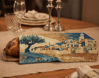 May include: A challah bread wrapped in a decorative cloth featuring a mosaic design of a tree and a village with blue rooftops. The scene is set on a table with candles, glassware, and silverware.