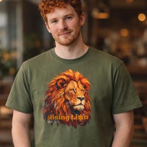 Könnte beinhalten: Olivgrünes T-Shirt mit einer detaillierten Illustration eines Löwenkopfes mit orangefarbener Mähne. Der Text "Rising Lion" und "Num 23:24" sind darunter gedruckt. Das Shirt besteht aus einem weichen Material.