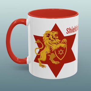 May include: White ceramic mug with a red interior and handle. The mug features a gold lion holding a shield with a Star of David on a red starburst. The word "Shield" is printed on the mug.