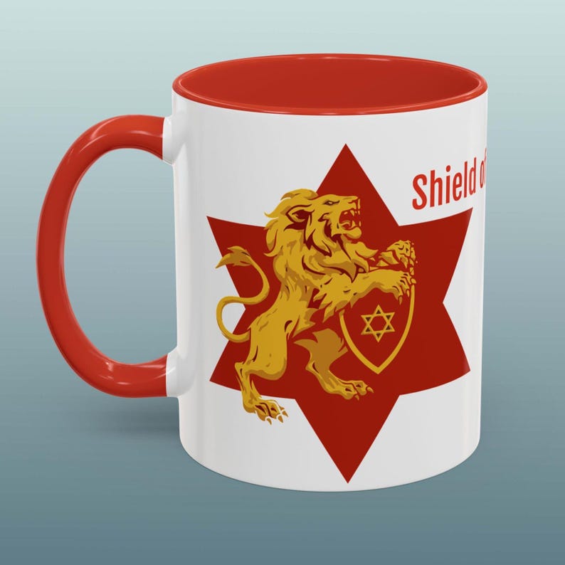 May include: White ceramic mug with a red interior and handle. The mug features a gold lion holding a shield with a Star of David on a red starburst. The word "Shield" is printed on the mug.