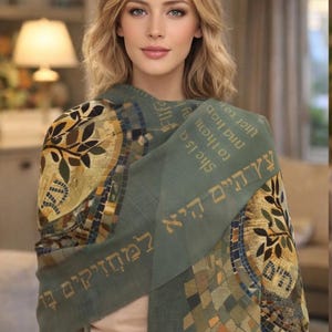 May include: A sage green scarf with mosaic and tree of life designs. The scarf has Hebrew text and is draped around the shoulders. The scarf is made of a soft material.