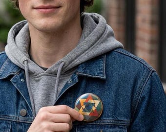 May include: A round button with a Star of David design in shades of orange, blue, green, and yellow. The button is held against a denim jacket. The Star of David is a symbol of Judaism.