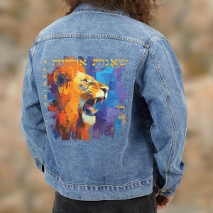 May include: A light blue denim jacket with a colorful lion graphic on the back. The lion is depicted with orange, yellow, and blue hues, with text above and to the side of the lion. The jacket has a classic design.