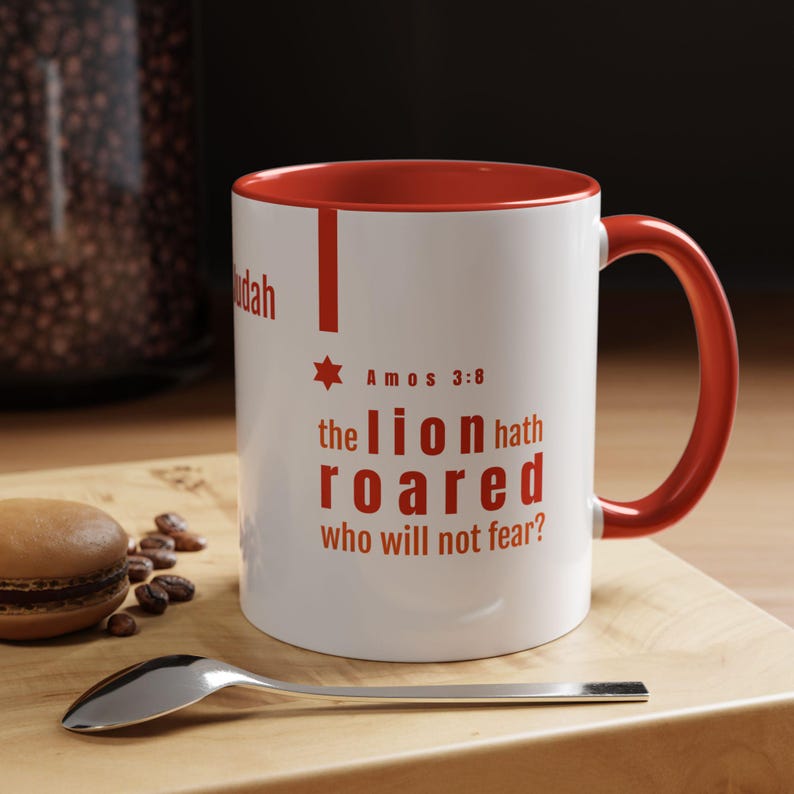 May include: White ceramic mug with a red interior and handle. The mug features red text that reads "the lion hath roared who will not fear?" with a star and the text "Amos 3:8". A spoon and a macaron are on a wooden surface.