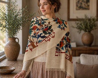 May include: A cream-colored shawl featuring a vibrant floral pattern in shades of red, green, and blue, accented with Hebrew text in red lettering. The shawl has a fringed hem and is draped over a light-colored sweater and a long, pleated skirt.